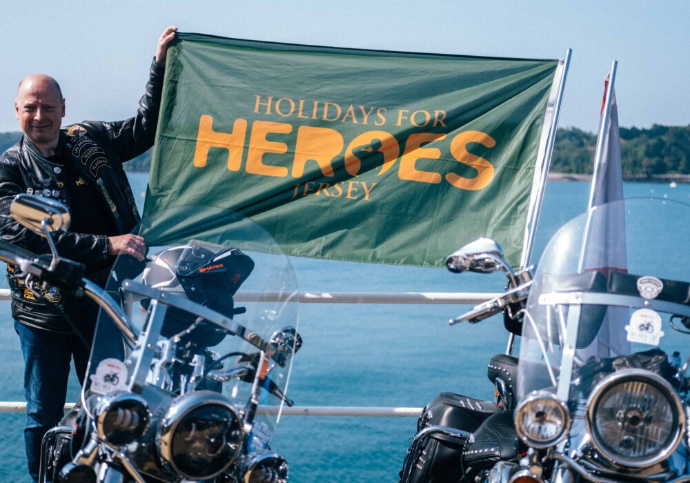 A motorcyclist holding a Holidays for Heroes Jersey banner next to bikes at a scenic spot, promoting the Big Ride Out 2024 sponsored by Islands Insurance.