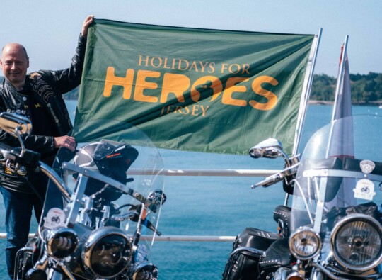 A motorcyclist holding a Holidays for Heroes Jersey banner next to bikes at a scenic spot, promoting the Big Ride Out 2024 sponsored by Islands Insurance.